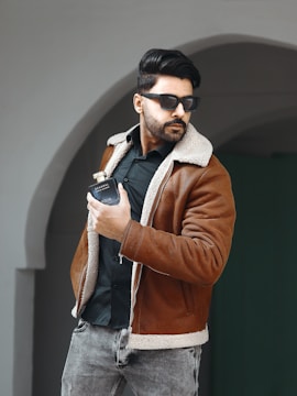 A man with dark hair and a beard is wearing sunglasses, a brown shearling jacket, and a black shirt. He is holding a bottle of cologne in his right hand. He stands in front of an archway, and his posture is slightly angled to the side.