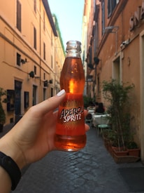 A hand holding a bottle of Aperol Spritz in a narrow, cobblestone alleyway. The buildings on either side are tall and rustic, painted in warm earthy tones. Potted plants and outdoor seating are visible, suggesting a cozy, European street setting. The lighting suggests a late afternoon or early evening ambiance.
