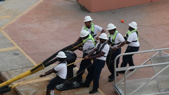 Five workers wearing white hard hats and reflective safety vests pull on a thick yellow and black rope. They are positioned on a concrete surface with a metal railing nearby. The workers appear focused and coordinated as they perform a task, likely related to maritime or industrial work.