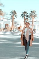 A stylish woman is walking outdoors in a sunny setting with palm trees and white buildings in the background. She is wearing a brown coat, headscarf, and beret, combined with a white top, jeans, and black boots.