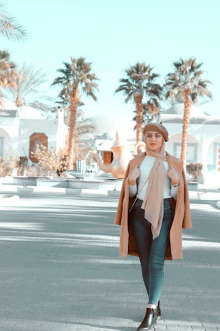 A stylish woman is walking outdoors in a sunny setting with palm trees and white buildings in the background. She is wearing a brown coat, headscarf, and beret, combined with a white top, jeans, and black boots.
