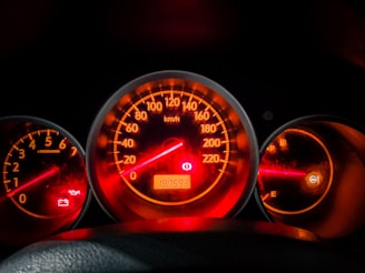 A car dashboard displaying several illuminated gauges, including a speedometer in the center showing a reading, a tachometer on the left, and a fuel gauge on the right. The background is dimly lit with red and orange highlights, indicating the dashboard lights are on. Warning lights for oil and battery are also visible.