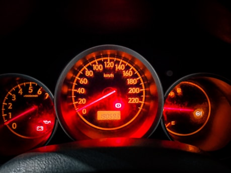 A car dashboard displaying several illuminated gauges, including a speedometer in the center showing a reading, a tachometer on the left, and a fuel gauge on the right. The background is dimly lit with red and orange highlights, indicating the dashboard lights are on. Warning lights for oil and battery are also visible.