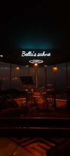 A dimly lit stage with music equipment including a drum set and a keyboard. There's a microphone stand in front of the instruments and a neon sign overhead that reads 'Bella's sahne'. The background shows large windows with blurred city lights visible through them.