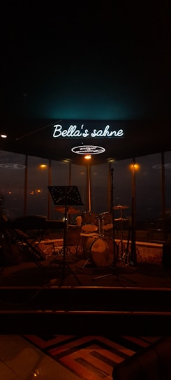 A dimly lit stage with music equipment including a drum set and a keyboard. There's a microphone stand in front of the instruments and a neon sign overhead that reads 'Bella's sahne'. The background shows large windows with blurred city lights visible through them.
