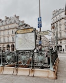 An ornate entrance to a metro station in a Parisian street, featuring decorative railings and a map of the metro network. Surrounding the entrance are historical buildings with classic French architecture. The atmosphere is overcast, and people are seen in the background.