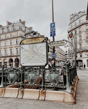 An ornate entrance to a metro station in a Parisian street, featuring decorative railings and a map of the metro network. Surrounding the entrance are historical buildings with classic French architecture. The atmosphere is overcast, and people are seen in the background.