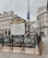 An ornate entrance to a metro station in a Parisian street, featuring decorative railings and a map of the metro network. Surrounding the entrance are historical buildings with classic French architecture. The atmosphere is overcast, and people are seen in the background.