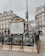 An ornate entrance to a metro station in a Parisian street, featuring decorative railings and a map of the metro network. Surrounding the entrance are historical buildings with classic French architecture. The atmosphere is overcast, and people are seen in the background.
