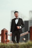A confident man wearing a classic black tuxedo with soft gold cufflinks standing beside a sleek black car.