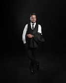 A man dressed in formal attire is standing confidently against a dark backdrop. He is wearing a white shirt, black vest, and tie with matching pants and shoes. One arm casually holds a black jacket over his shoulder.