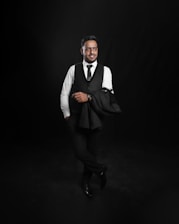 A man dressed in formal attire is standing confidently against a dark backdrop. He is wearing a white shirt, black vest, and tie with matching pants and shoes. One arm casually holds a black jacket over his shoulder.