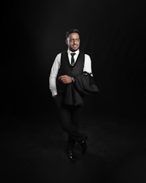 A man dressed in formal attire is standing confidently against a dark backdrop. He is wearing a white shirt, black vest, and tie with matching pants and shoes. One arm casually holds a black jacket over his shoulder.
