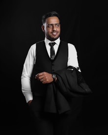 A man in formal attire, including a black vest and tie over a white shirt, holds a suit jacket draped over his arm. He wears a watch on his left wrist and displays a slight smile while standing against a dark background.