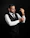 A man in formal attire appears to be adjusting his cufflinks. He is wearing a black vest over a white dress shirt, a black tie, and matching trousers. The background is solid black, creating a dramatic contrast with his outfit.