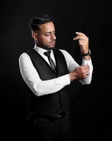 A man in formal attire appears to be adjusting his cufflinks. He is wearing a black vest over a white dress shirt, a black tie, and matching trousers. The background is solid black, creating a dramatic contrast with his outfit.