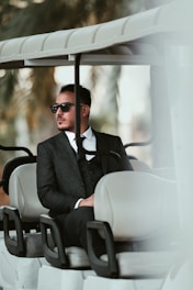 A man wearing a dark suit and sunglasses is seated in a golf cart. The setting has a tropical feel with blurred palm leaves in the background, suggesting a warm, leisurely environment.
