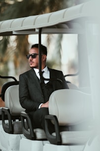 A man wearing a dark suit and sunglasses is seated in a golf cart. The setting has a tropical feel with blurred palm leaves in the background, suggesting a warm, leisurely environment.