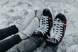 A pair of black hockey skates with orange accents and white figure skates rest side by side on an ice surface. The skates are worn by individuals dressed in black clothing, with the snow-covered ice creating a wintry backdrop.