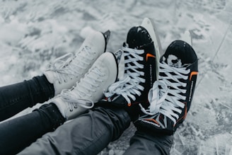 A pair of black hockey skates with orange accents and white figure skates rest side by side on an ice surface. The skates are worn by individuals dressed in black clothing, with the snow-covered ice creating a wintry backdrop.