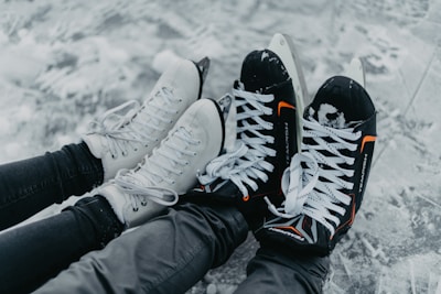 A pair of black hockey skates with orange accents and white figure skates rest side by side on an ice surface. The skates are worn by individuals dressed in black clothing, with the snow-covered ice creating a wintry backdrop.