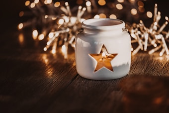 A white ceramic candle holder with a star-shaped cutout sits on a wooden surface. Inside, a lit candle casts a warm glow through the star opening. In the background, there are blurred twinkling lights, creating a cozy and festive atmosphere.