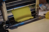 A sheet of yellow dough is being rolled through a metal pasta machine, with a smooth surface and precise, straight edges. The machine is shiny and metallic, with rollers that flatten the dough evenly. The scene conveys a process of making fresh pasta or another type of dough-based food product.