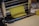 A sheet of yellow dough is being rolled through a metal pasta machine, with a smooth surface and precise, straight edges. The machine is shiny and metallic, with rollers that flatten the dough evenly. The scene conveys a process of making fresh pasta or another type of dough-based food product.
