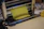 A sheet of yellow dough is being rolled through a metal pasta machine, with a smooth surface and precise, straight edges. The machine is shiny and metallic, with rollers that flatten the dough evenly. The scene conveys a process of making fresh pasta or another type of dough-based food product.