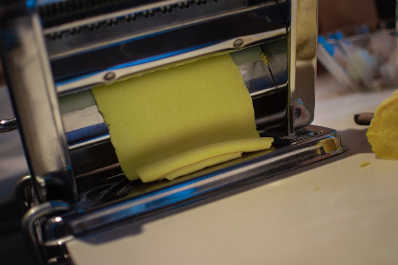 A sheet of yellow dough is being rolled through a metal pasta machine, with a smooth surface and precise, straight edges. The machine is shiny and metallic, with rollers that flatten the dough evenly. The scene conveys a process of making fresh pasta or another type of dough-based food product.