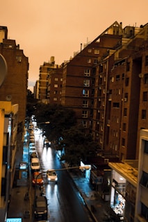 A city street scene during twilight with several multi-story brick buildings on both sides. The street is wet, likely from recent rain, reflecting the lights of street lamps and car headlights. There are parked cars along the sides of the street and trees lining the road. Some sklep signs are visible at the ground level of the buildings.