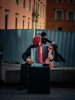 A street musician sits on a stone bench playing an accordion. The musician wears a red and black checkered hat and is dressed in dark clothing. The background features a large metal fence and an old building with a pink and brown facade.