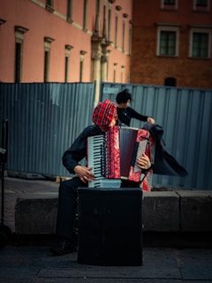 A street musician sits on a stone bench playing an accordion. The musician wears a red and black checkered hat and is dressed in dark clothing. The background features a large metal fence and an old building with a pink and brown facade.