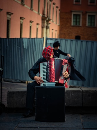 A street musician sits on a stone bench playing an accordion. The musician wears a red and black checkered hat and is dressed in dark clothing. The background features a large metal fence and an old building with a pink and brown facade.