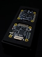 Close-up view of an electronic circuit board encased in a black rectangular container. The visible components include microchips, a USB port, and other small electronic parts. Yellow circular fixtures can be seen securing parts of the device.