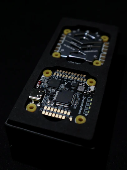 Close-up view of an electronic circuit board encased in a black rectangular container. The visible components include microchips, a USB port, and other small electronic parts. Yellow circular fixtures can be seen securing parts of the device.