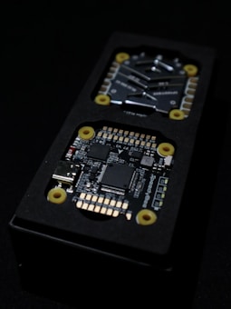 Close-up view of an electronic circuit board encased in a black rectangular container. The visible components include microchips, a USB port, and other small electronic parts. Yellow circular fixtures can be seen securing parts of the device.