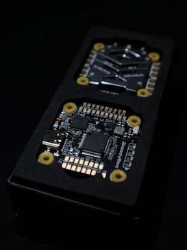 Close-up view of an electronic circuit board encased in a black rectangular container. The visible components include microchips, a USB port, and other small electronic parts. Yellow circular fixtures can be seen securing parts of the device.