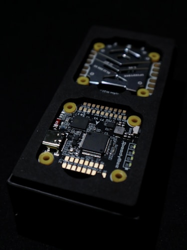 Close-up view of an electronic circuit board encased in a black rectangular container. The visible components include microchips, a USB port, and other small electronic parts. Yellow circular fixtures can be seen securing parts of the device.