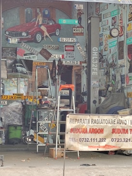 A cluttered workshop space filled with various car license plates, signs, and vintage posters. A red classic car painted on the wall has a pin-up model sitting on it. A ladder is placed in the middle of the space, surrounded by tools and mechanical parts. A banner in the foreground advertises auto and radiator repair services, listing contact numbers. The overall atmosphere suggests automotive workshop or restoration.