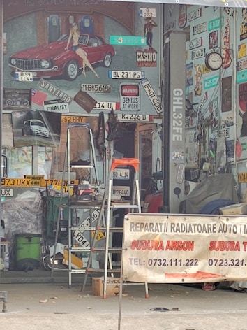 A cluttered workshop space filled with various car license plates, signs, and vintage posters. A red classic car painted on the wall has a pin-up model sitting on it. A ladder is placed in the middle of the space, surrounded by tools and mechanical parts. A banner in the foreground advertises auto and radiator repair services, listing contact numbers. The overall atmosphere suggests automotive workshop or restoration.
