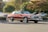 A classic, red vintage car is parked in an outdoor parking lot. The car has distinctive features typical of older models, such as a boxy shape, chrome detailing, and prominent grille. Surrounding the car are several other modern vehicles in a lightly wooded area with green trees in the background, providing contrast between the old and new.