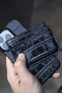 A close-up view of a hand holding a sleek, black crocodile-patterned leather cardholder with the brand name 'GENTCREATE' embossed in gold. The cardholder is positioned alongside a smartphone with a matching black leather phone case.