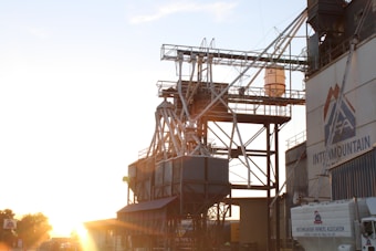 An industrial structure with metal framework and pipes is silhouetted against a bright setting sun. To the right, a building with the logo of an association is visible, along with a parked truck.