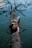 A person lies on their back on a fallen tree trunk that extends over a calm body of water. Branches protrude in various directions, and the person's arms are extended outward, reaching towards the camera. The scene conveys a sense of peace and connection with nature.