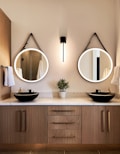 A modern bathroom scene featuring a wooden vanity with two round mirrors, each with a black strap hanging above. A sleek wall light fixture is centered between the mirrors. On the marble countertop, there are two black vessel sinks with gold faucets, flanked by white hand towels. A small potted plant is placed between the sinks.