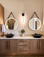 A modern bathroom scene featuring a wooden vanity with two round mirrors, each with a black strap hanging above. A sleek wall light fixture is centered between the mirrors. On the marble countertop, there are two black vessel sinks with gold faucets, flanked by white hand towels. A small potted plant is placed between the sinks.