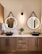 A modern bathroom scene featuring a wooden vanity with two round mirrors, each with a black strap hanging above. A sleek wall light fixture is centered between the mirrors. On the marble countertop, there are two black vessel sinks with gold faucets, flanked by white hand towels. A small potted plant is placed between the sinks.