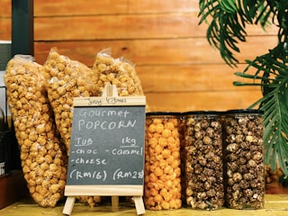 Several packages and jars of gourmet popcorn are displayed on a wooden surface. The flavors include chocolate, caramel, and cheese, as indicated by a small chalkboard. A plant decorates the right side of the image.