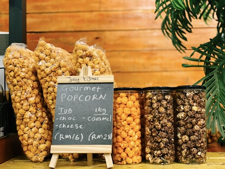 Several packages and jars of gourmet popcorn are displayed on a wooden surface. The flavors include chocolate, caramel, and cheese, as indicated by a small chalkboard. A plant decorates the right side of the image.